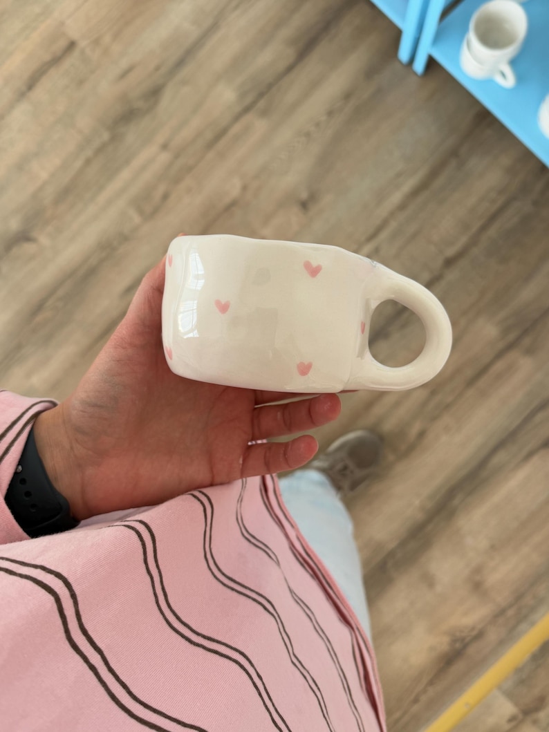 Peut inclure: Une tasse en c&eacute;ramique blanche avec une anse arrondie et des motifs en forme de c&oelig;ur roses. La tasse est tenue dans la main d'une personne. L'arri&egrave;re-plan comprend un sol en bois et un aper&ccedil;u d'une &eacute;tag&egrave;re bleue.