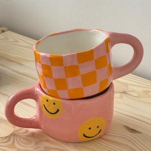 May include: Two ceramic mugs stacked on top of each other. The top mug has a pink and orange checkerboard pattern. The bottom mug is pink with two yellow smiley faces. Both mugs have pink handles.