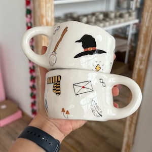 Puede incluir: Dos tazas de cerámica blancas con ilustraciones negras de un sombrero de bruja, una escoba, una bufanda, un sobre, gafas y un rayo. Las tazas tienen asas grandes y están apiladas.