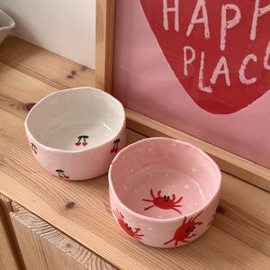 Peut inclure: Deux petits bols en céramique faits à la main, de forme ronde avec des bords irréguliers et ondulés. Un bol est rose clair avec des cerises rouges à l'intérieur et à l'extérieur. L'autre est rose clair à pois blancs avec des crabes rouges.
