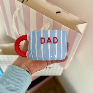 Peut inclure: Une tasse à rayures bleues et blanches avec le mot "DAD" en lettres rouges. La tasse a une anse rouge et est tenue dans une main. Un sac cadeau à rayures roses et blanches est en arrière-plan.