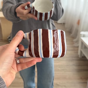 May include: Two ceramic mugs with brown, white, and light blue vertical stripes. The mugs have a unique, irregular shape and a handle. One mug is held up, while the other is held in the foreground.