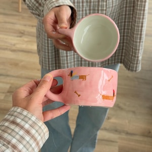 Op de afbeelding: Roze keramische mok met een handvat, versierd met teckel- en botmotieven. De binnenkant is wit met een roze rand. Een andere roze mok met een handvat wordt erboven gehouden.