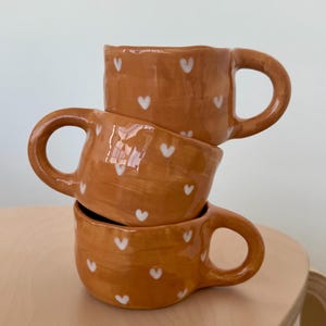 Op de afbeelding: Drie gestapelde keramische mokken in een warme oranje tint, versierd met kleine witte hartpatronen. Elke mok heeft een afgeronde handgreep. De mokken zijn op elkaar gestapeld, wat een visueel aantrekkelijke opstelling creëert. De mokken staan op een lichtbruin houten oppervlak.