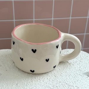 Può includere: Una piccola tazza in ceramica bianco sporco con bordo e manico rosa. La tazza è decorata con piccole forme di cuore nere. La tazza è appoggiata su una superficie bianca.