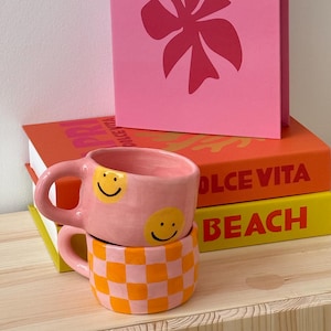 May include: Two ceramic mugs stacked on a wooden surface. The top mug is pink with yellow smiley faces. The bottom mug has a pink and orange checkerboard pattern. A pink book with a floral design and the word "BEACH" is in the background.