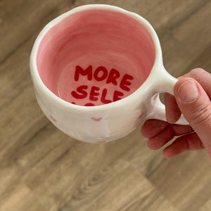 Peut inclure: Tasse en c&eacute;ramique blanche avec int&eacute;rieur et anse roses. Les mots "MORE SELF" sont &eacute;crits en rouge &agrave; l'int&eacute;rieur de la tasse. De petits c&oelig;urs roses sont peints &agrave; l'ext&eacute;rieur de la tasse.