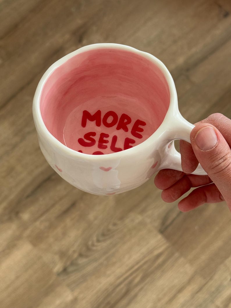 Peut inclure: Tasse en c&eacute;ramique blanche avec int&eacute;rieur et anse roses. Les mots "MORE SELF" sont &eacute;crits en rouge &agrave; l'int&eacute;rieur de la tasse. De petits c&oelig;urs roses sont peints &agrave; l'ext&eacute;rieur de la tasse.