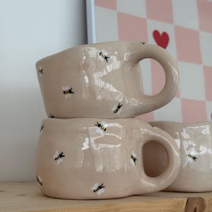 May include: Two beige ceramic mugs stacked on a wooden shelf. Each mug has a large handle and is decorated with small, hand-painted bee designs. The background includes a pink and white checkered pattern with red hearts.