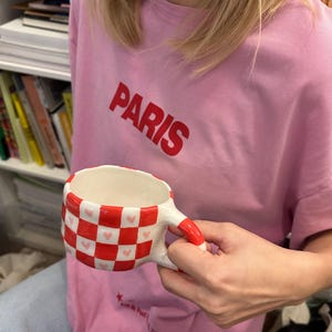 Pode incluir: Uma caneca de cerâmica com um padrão xadrez vermelho e branco, acentuada com pequenos corações rosa. A alça e a borda são vermelhas. A caneca é segurada em frente a uma camiseta rosa com a palavra "PARIS" impressa em vermelho.