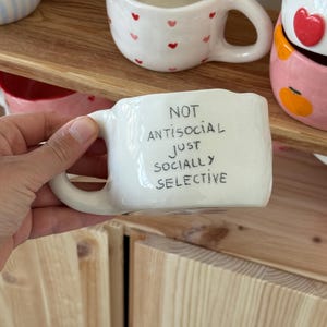 Könnte beinhalten: Weiße Keramiktasse mit dem Text "NOT ANTISOCIAL JUST SOCIALLY SELECTIVE" in schwarzer Schrift. Die Tasse hat einen gebogenen Henkel. Weitere Tassen mit Herz- und Fruchtdesigns sind im Hintergrund zu sehen.
