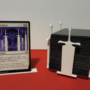 Puede incluir: Una carta de Magic: The Gathering titulada "Ghostly Prison" con una ilustración púrpura de una criatura tras las rejas. Un tarjetero blanco y un expositor de cartas blanco también están en la imagen. El tarjetero tiene un diseño de espada.
