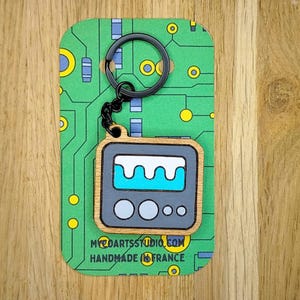Peut inclure: Un porte-clés en bois en forme de télévision rétro avec un cadre gris foncé, un écran affichant un motif de vague bleu clair et deux boutons circulaires. Le porte-clés est fixé à un anneau en métal noir sur une carte à motif de circuit imprimé.