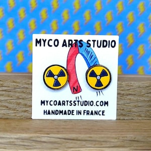 Op de afbeelding: Een paar gele en zwarte oorbellen met een stralingssymbool. De oorbellen liggen op een witte kaart met de tekst "MYCO ARTS STUDIO" en "MYCOARTSSTUDIO.COM HANDMADE IN FRANCE". De achtergrond is een blauw patroon met gele bliksemschichten.