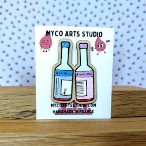 Puede incluir: Pin de madera con dos botellas estilizadas con las etiquetas "Aeróbico" y "Anaeróbico" con detalles en azul y rosa. El pin está sobre una tarjeta blanca con el texto "MYCO ARTS STUDIO" y "HANDMADE IN FRANCE".