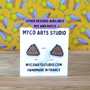 Può includere: Orecchini a forma di cacca marrone con faccine sorridenti, esposti su una carta bianca. La carta presenta il testo "MYCO ARTS STUDIO" e "HANDMADE IN FRANCE". Lo sfondo è un motivo giallo con stelle.