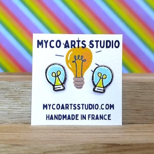 Può includere: Una carta bianca con il testo "MYCO ARTS STUDIO" e "MYCOARTSSTUDIO.COM HANDMADE IN FRANCE" presenta tre illustrazioni di lampadine. La lampadina centrale è arancione, mentre quelle laterali sono blu. Lo sfondo è un motivo arcobaleno.