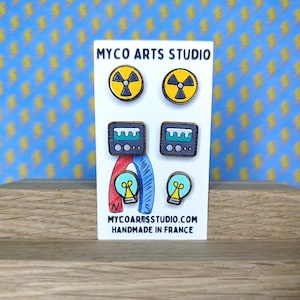 Op de afbeelding: Een witte kaart toont zes houten oorstekers met wetenschappelijke ontwerpen. De ontwerpen omvatten een geel stralingssymbool, een grijs en blauw apparaat en een gloeilamp. De kaart heeft de tekst "MYCO ARTS STUDIO" en "HANDMADE IN FRANCE".