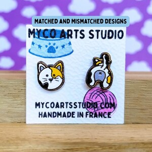 Può includere: Un paio di orecchini a perno in legno raffiguranti il muso di un gatto calico e il posteriore di un gatto calico. Gli orecchini sono esposti su un cartoncino bianco con la scritta MYCO ARTS STUDIO, MATCHED AND MISMATCHED DESIGNS, MYCOARTSSTUDIO.COM, HANDMADE IN FRANCE.