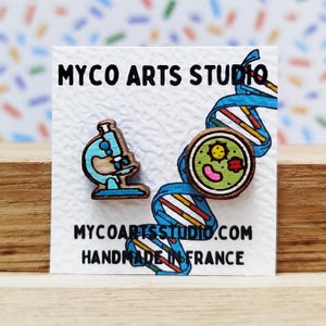 Op de afbeelding: Een paar houten oorbellen met een microscoop en een cel met een glimlachend gezicht. De oorbellen bevinden zich op een witte kaart met de tekst "MYCO ARTS STUDIO.COM HANDMADE IN FRANCE".