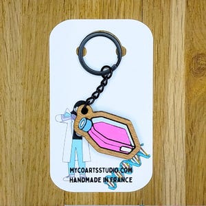 Può includere: Un portachiavi in legno con un ciondolo a forma di fiaschetta da laboratorio riempita di liquido rosa e un tappo blu, fissato a un anello in metallo nero. Il cartoncino mostra l'illustrazione di uno scienziato, un filamento di DNA e il testo MYCOARTSSTUDIO.COM HANDMADE IN FRANCE.
