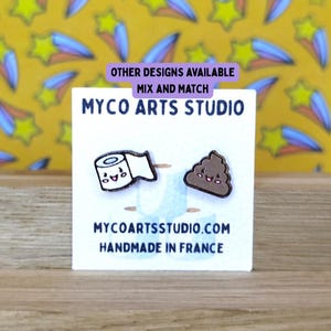 Può includere: Una carta bianca con il testo "MYCO ARTS STUDIO" e "MYCOARTSSTUDIO.COM HANDMADE IN FRANCE". La carta presenta due piccole spille in stile cartone animato: un rotolo di carta igienica e un'emoji di cacca sorridente. Lo sfondo è un motivo colorato.