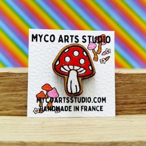 Può includere: Uno spillo di legno a forma di fungo con un cappello rosso e macchie bianche, un gambo bianco e una base marrone. Lo spillo è su una carta bianca con il testo "MYCO ARTS STUDIO.COM HANDMADE IN FRANCE".
