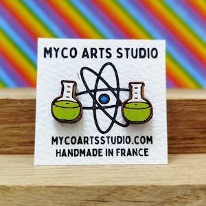 Może przedstawiać: Para drewnianych kolczyków w kształcie zlewki z zielonym płynem w środku. Kolczyki znajdują się na białej kartce z tekstem "MYCO ARTS STUDIO" i "MYCOARTSTUDIO.COM HANDMADE IN FRANCE".