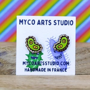 Könnte beinhalten: Ein Paar grüne und gelbe Holzohrringe in Form von Bakterien. Die Ohrringe befinden sich auf einer weißen Karte mit dem Text "Myco Arts Studio" und "Handgefertigt in Frankreich".