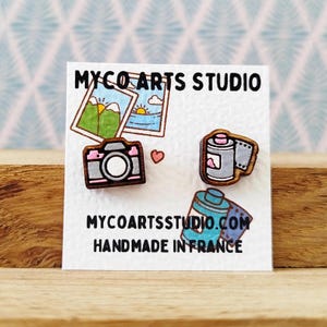 May include: Wooden earrings featuring a camera, film roll, and a film canister. The earrings are painted in blue, pink, and brown. The card reads "Mycoartsstudio.com Handmade in France".