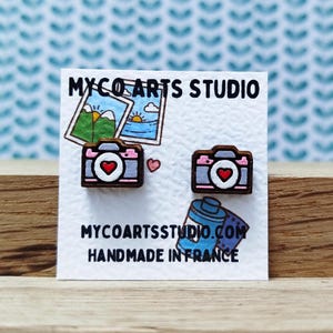 May include: A pair of wooden camera earrings with a pink heart detail. The earrings are on a white card with the text "MYCOARTSTUDIO.COM HANDMADE IN FRANCE".