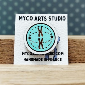 Può includere: Uno spillo di legno con un cerchio blu e due X nere all'interno. Il cerchio ha un bordo bianco e una linea punteggiata attorno al bordo. Lo spillo dice "Myco Arts Studio" e "Fatto a mano in Francia".