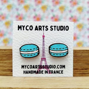 Könnte beinhalten: Ein Paar Holzohrringe in Form von blauen Macarons mit weißer Füllung. Die Ohrringe werden auf einer weißen Karte mit dem Text "Myco Arts Studio" und "Handgefertigt in Frankreich" präsentiert.