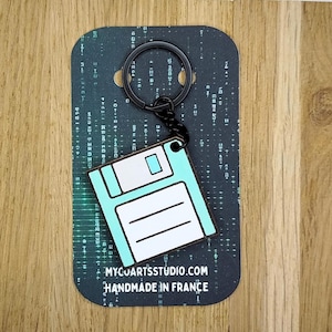 Puede incluir: Un llavero de disquete en color azul claro y blanco unido a una anilla de metal negro. El llavero está montado sobre una tarjeta rectangular con un patrón de matriz digital verde oscuro, el texto MYCUARTSSTUDIO.COM y HANDMADE IN FRANCE.