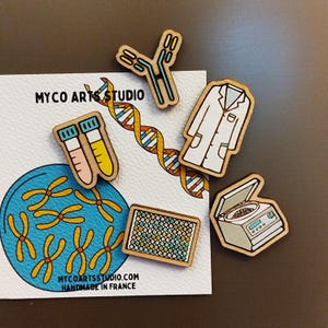May include: A set of wooden science-themed pins featuring a lab coat, test tubes, a centrifuge, a DNA strand, a petri dish with chromosomes, and a well plate. The pins are handmade in France and are sold by MyCoArtStudio.com.