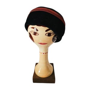 Può includere: Un cappello cloche nero con una fascia rosso scuro, esposto su una testa decorativa in ceramica. La testa è dipinta con tratti del viso e poggia su una base di legno. Il cappello è in stile vintage.