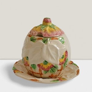 May include: Cream-colored ceramic serving dish with a lid and matching saucer. The dish is decorated with embossed leaves and colorful floral accents in yellow, pink, and green. The lid has a small handle.
