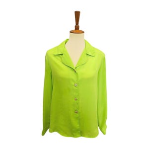 May include: A lime green long-sleeve blouse with a collared neckline and button-down front. The buttons are embellished with a decorative element. The blouse is displayed on a mannequin against a white background. The fabric has a slight sheen.