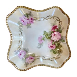 May include: A square, scalloped-edge dish with a cream-colored background. The dish is decorated with pink roses and green leaves, with a gold trim around the edge. The roses are painted in a realistic style, and the dish appears to be made of porcelain.