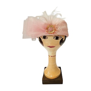 May include: A pink straw hat with feather and floral embellishments, displayed on a painted wooden mannequin head. The hat has a layered veil and a beaded floral accent, suitable for vintage-inspired fashion. The mannequin is on a wooden stand.