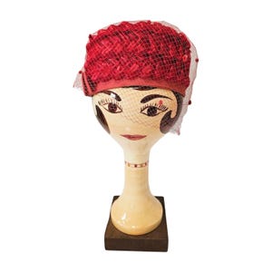 Puede incluir: Un sombrero rojo vintage con velo de red, exhibido en una cabeza de maniquí decorativa. El sombrero presenta una corona texturizada y una banda de satén. La cabeza del maniquí está pintada con rasgos faciales y descansa sobre una base de madera.