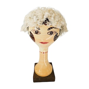 May include: A vintage-style mannequin head with a painted face, wearing a white hat with a veil. The hat is adorned with ruffled fabric. The mannequin is mounted on a wooden base, showcasing a retro aesthetic.
