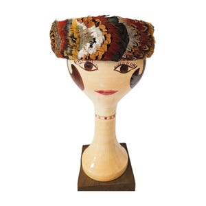 Puede incluir: Un sombrero decorativo con un diseño de plumas coloridas, exhibido en una cabeza de maniquí. El sombrero presenta una mezcla de plumas marrones, naranjas, blancas y azules. La cabeza del maniquí está pintada con rasgos faciales y se asienta sobre una base de madera.