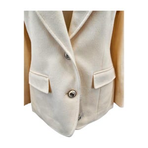 Pode incluir: Blazer creme com um design clássico. O blazer apresenta lapelas entalhadas, dois bolsos frontais e botões decorativos. O tecido parece ser uma mistura de lã macia. O blazer é de cor neutra, adequado para várias roupas.