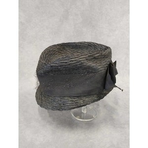 May include: A vintage-style black hat with a textured, woven appearance. The hat features a wide, dark band and a decorative bow on the side. The hat is displayed on a clear stand, showcasing its shape and design.