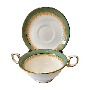 Puede incluir: Un juego de taza y platillo blanco y verde. La taza tiene dos asas y un borde dorado. El platillo y la taza tienen un borde verde con un diseño floral dorado. El conjunto está sobre un fondo blanco.