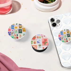 May include: Two phone grips with a colorful collage of vintage postage stamps featuring flowers and other designs. The phone grips are white with a black base. The phone grip on the left is on a white surface. The phone grip on the right is on a white phone case with a blue seashell pattern.