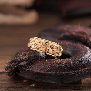 May include: A gold-colored ring with an eye pattern. The ring is made of a shiny metal and has a detailed design of stylized eyes around the band. The ring is displayed on a dark, textured surface, suggesting a jewelry or accessory theme.