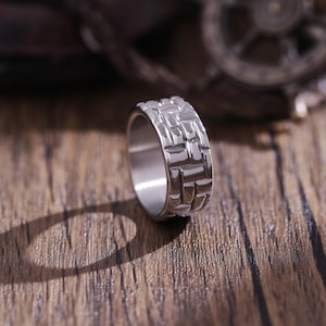 Könnte beinhalten: Ein silberner Ring mit einem strukturierten, ziegelartigen Muster. Der Ring liegt auf einer Holzoberfläche.