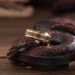 May include: A gold ring with a black braided design and runes inscribed around the band. The ring is resting on a dark brown surface.